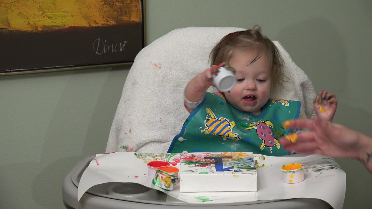 Baby in a high chair holds a small paint cup over a painted tray.