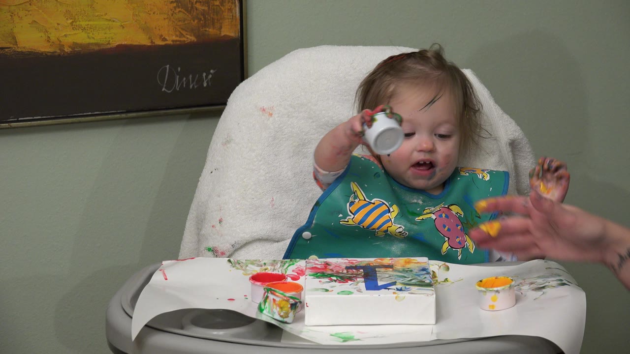 In Westminster, Colorado, the baby keeps one paint cup raised as an adult hand enters near the painted tray and open paint containers.