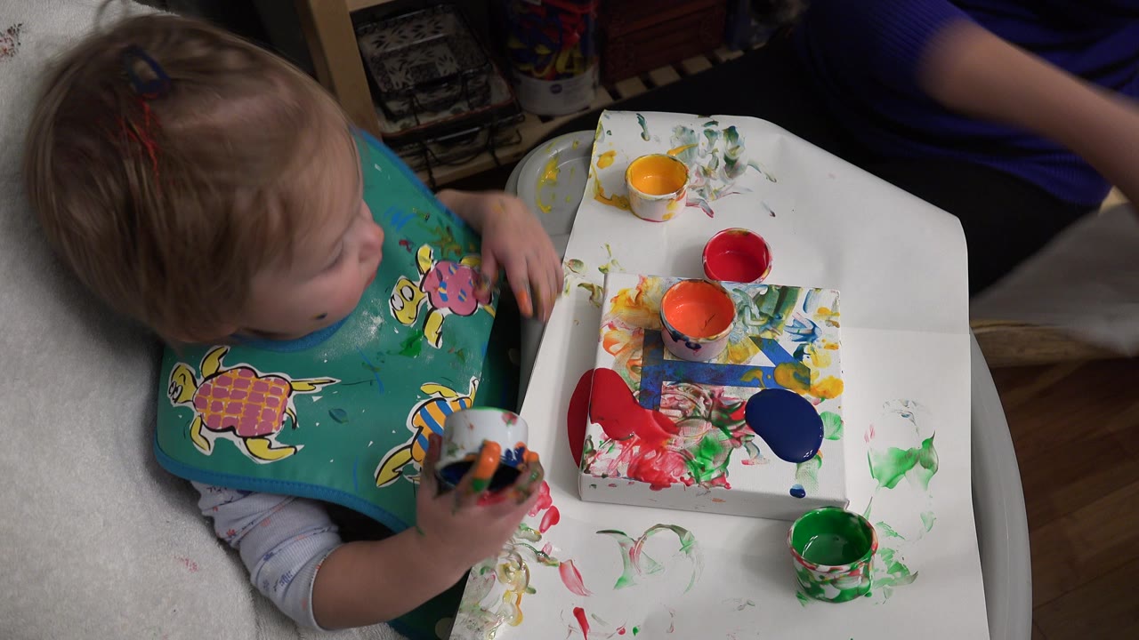 Toddler in a high chair holding a paint cup beside a messy tray.