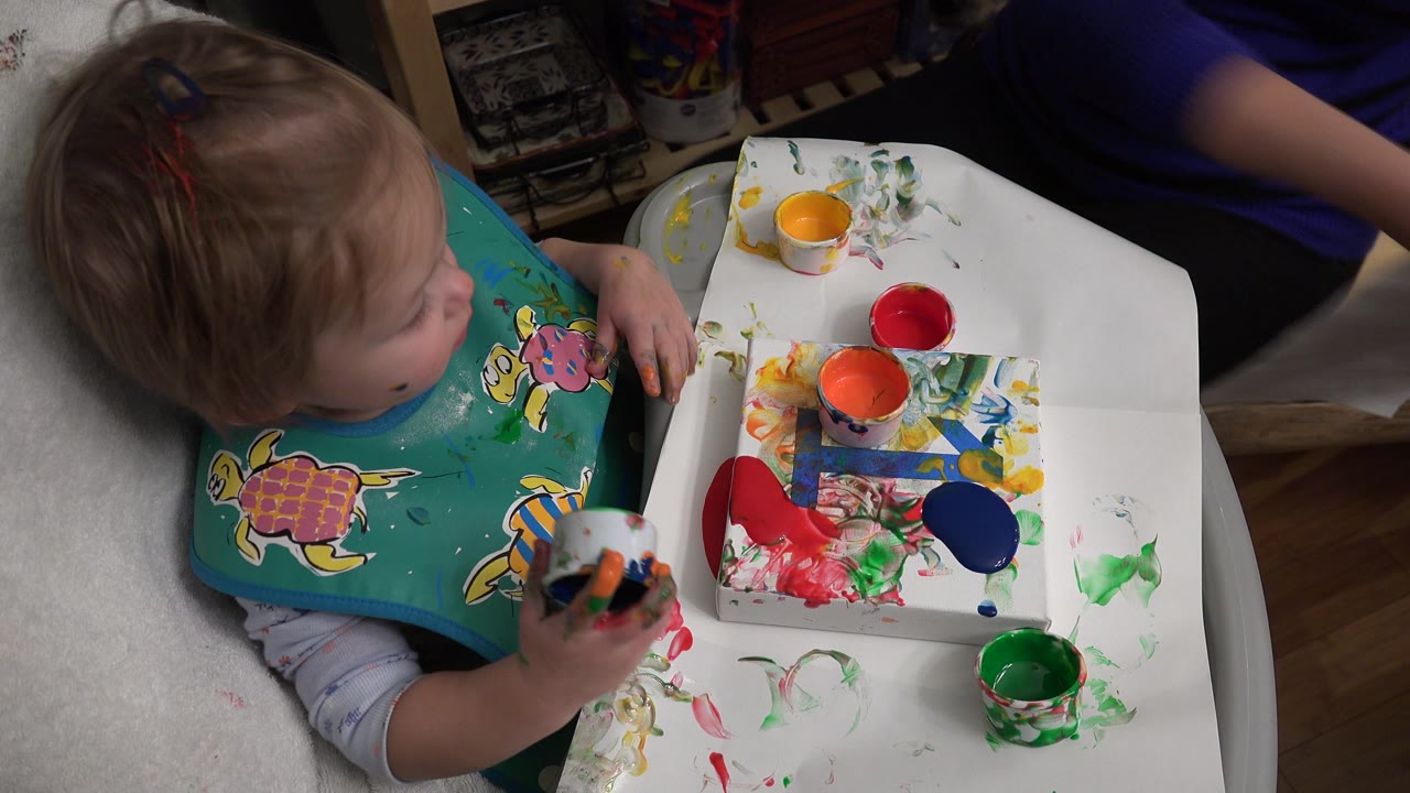 In Westminster, Colorado, the toddler turns toward the paint tray while holding a small cup near red, blue, green, yellow, and orange paint marks.