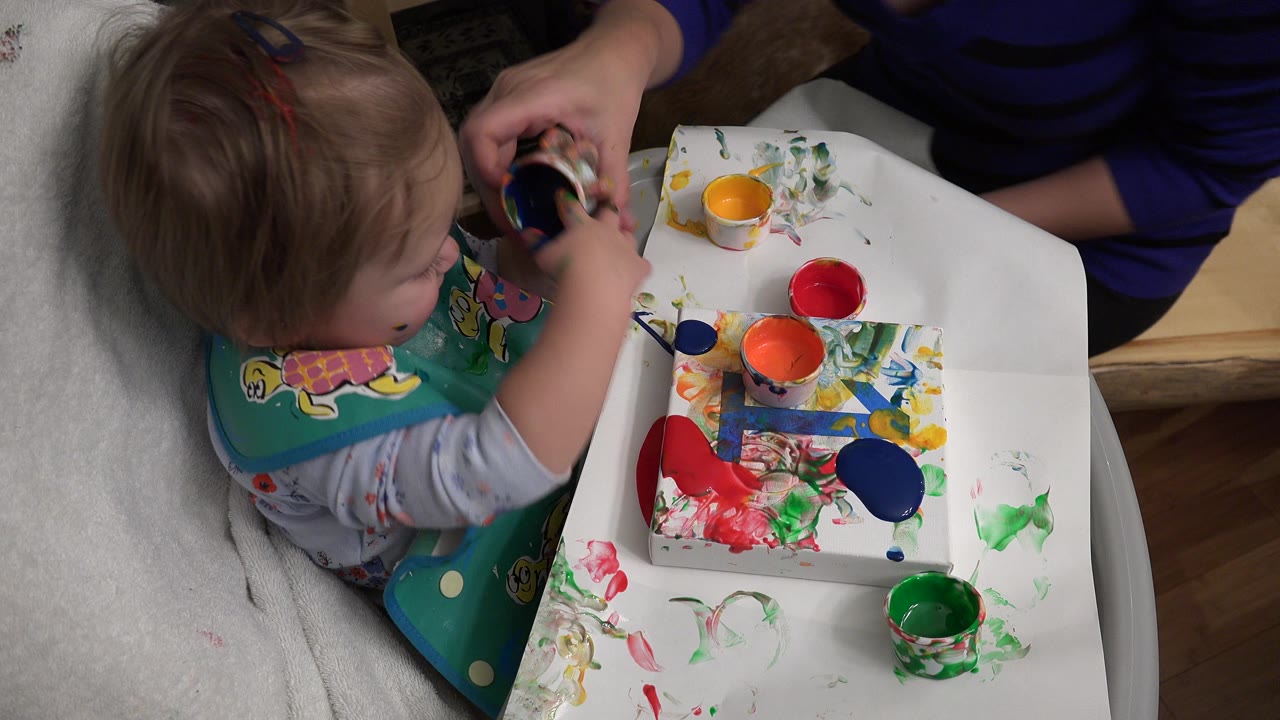 Toddler finger painting at a white tray table with an adult nearby.