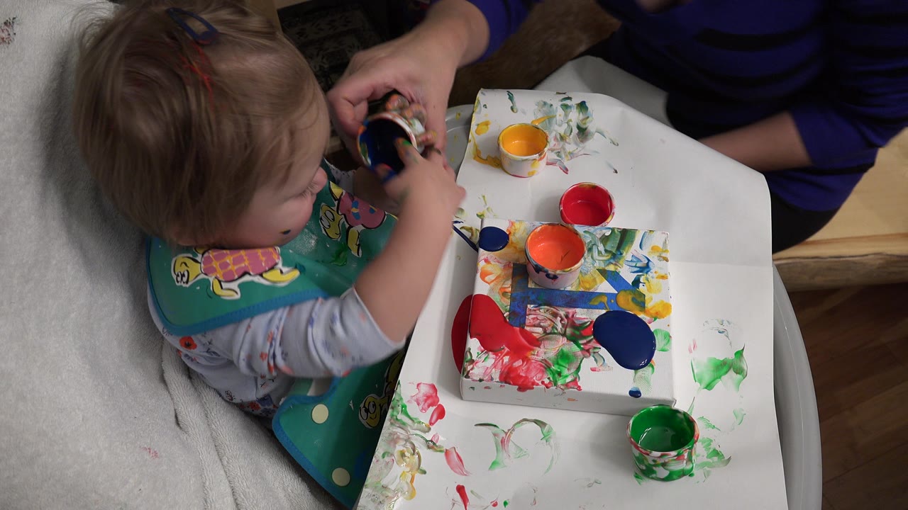 In Westminster, Colorado, the child holds a small paint container above paper marked with red, blue, green, and orange paint while the adult remains cropped beside the table.