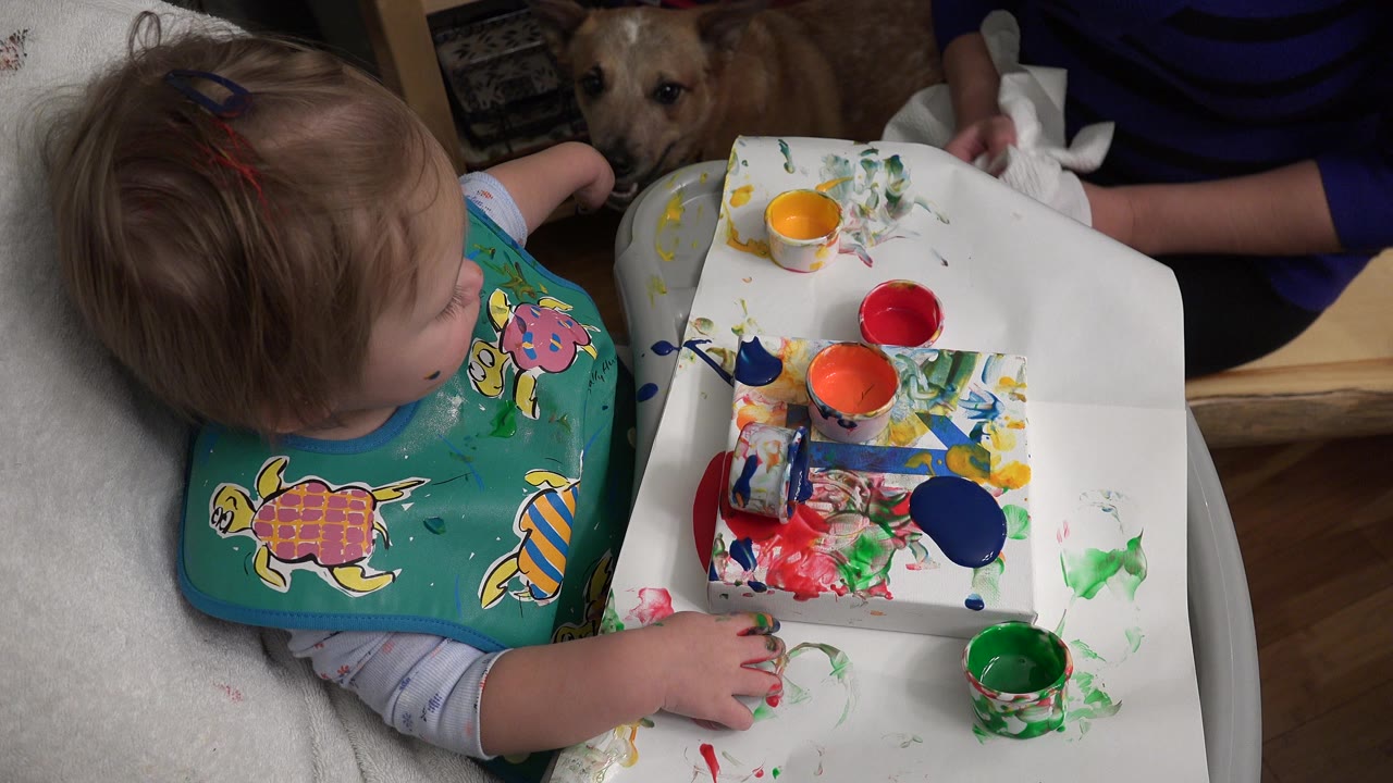 Toddler in a high chair reaches toward finger paint on a tray.