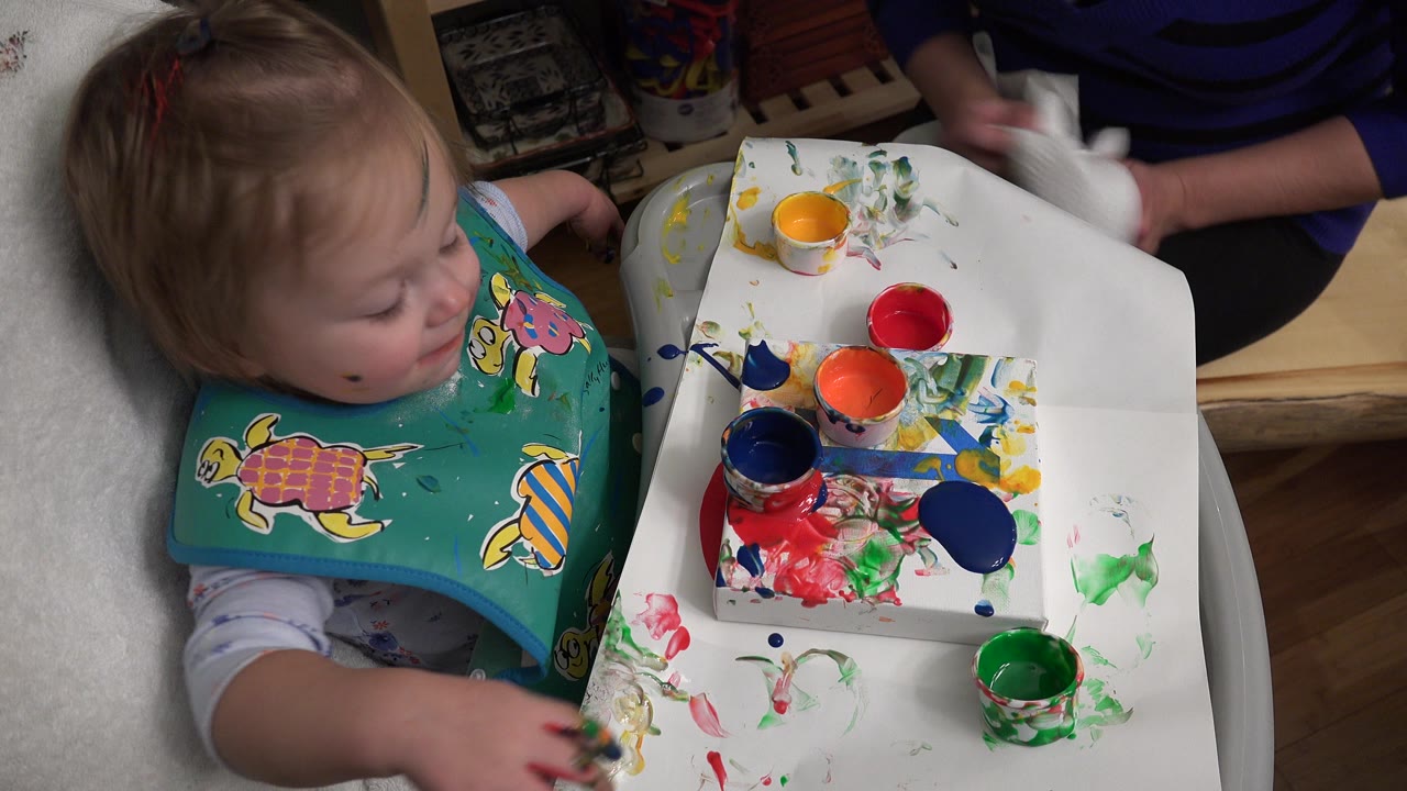A toddler in a green turtle bib sits at a white tray with red, blue, green, and orange paint cups during a Westminster, Colorado home art moment.