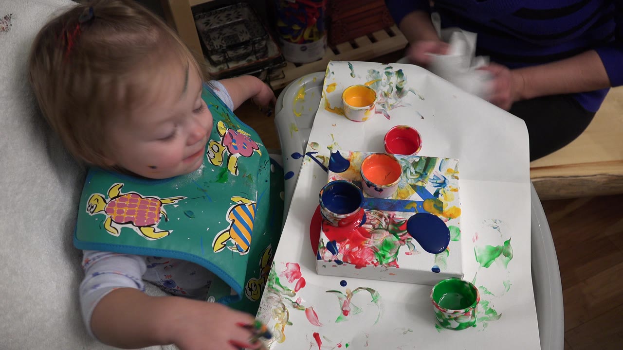 The child remains beside the white tray while an adult hand at the top right handles paper near the colorful paint cups in Westminster, Colorado.