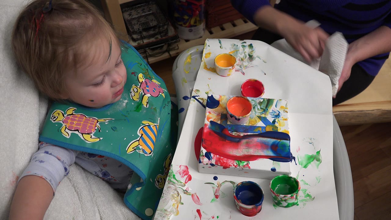 Baby in a green bib beside a white tray of paint cups.