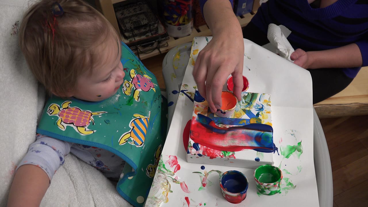 An adult hand reaches toward an orange paint cup on the tray while the baby sits close by in Westminster, Colorado.