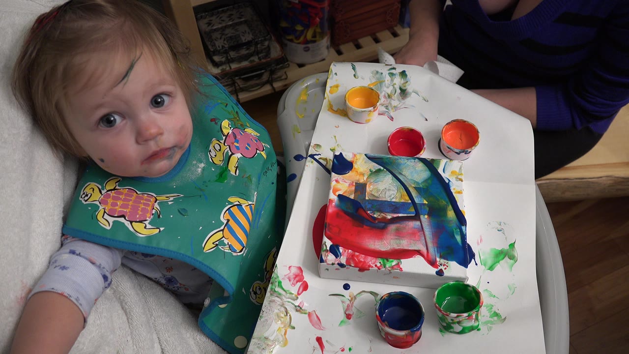 Infant in a paint smock beside finger paint cups on a highchair tray.