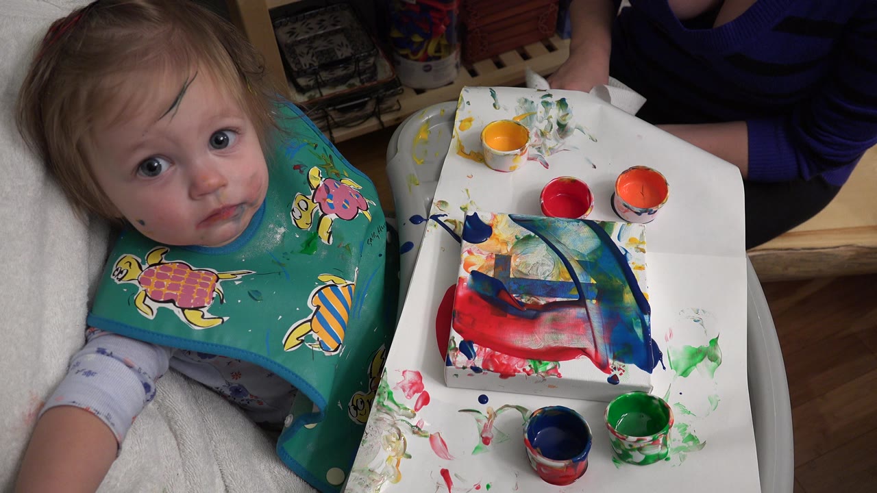 In Westminster, Colorado, the infant faces the camera while red, orange, green, blue, and yellow paint cups sit around a small painted surface on the tray.
