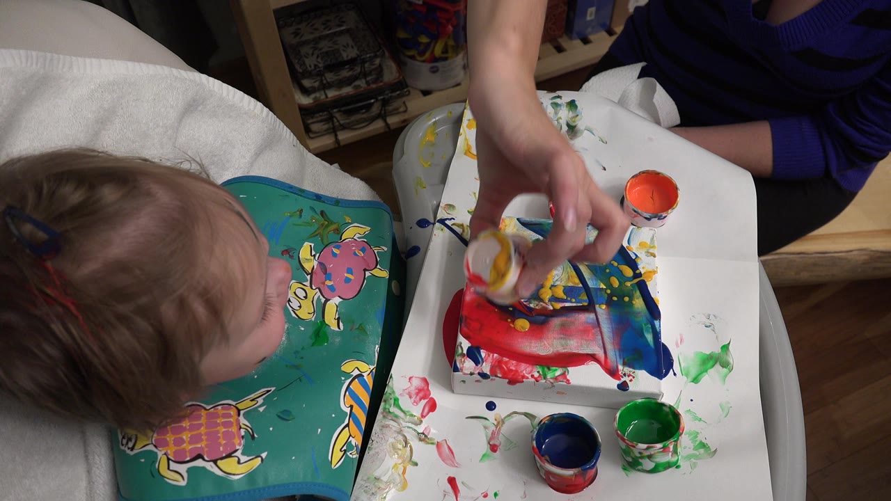 In Westminster, Colorado, the toddler leans toward the tray as the adult hand applies paint to a small paper or craft surface near open paint cups.