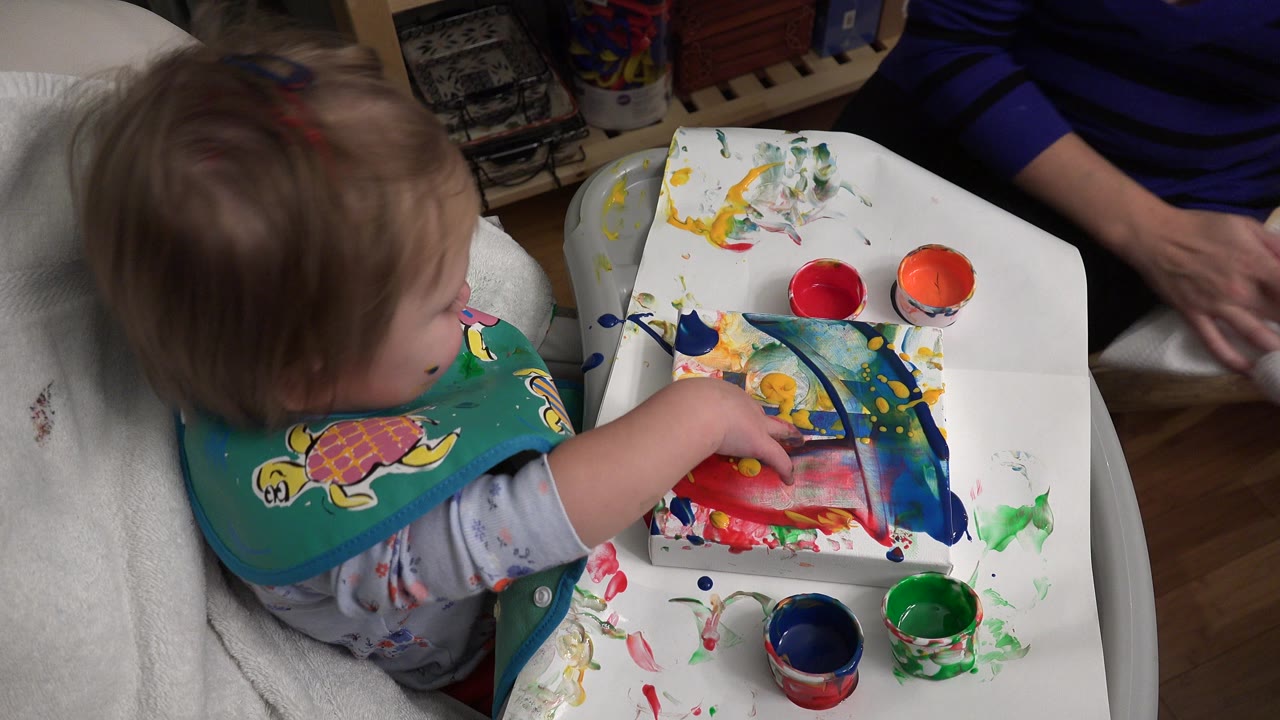 Toddler in a bib sits beside a paint-covered tray with colored cups.