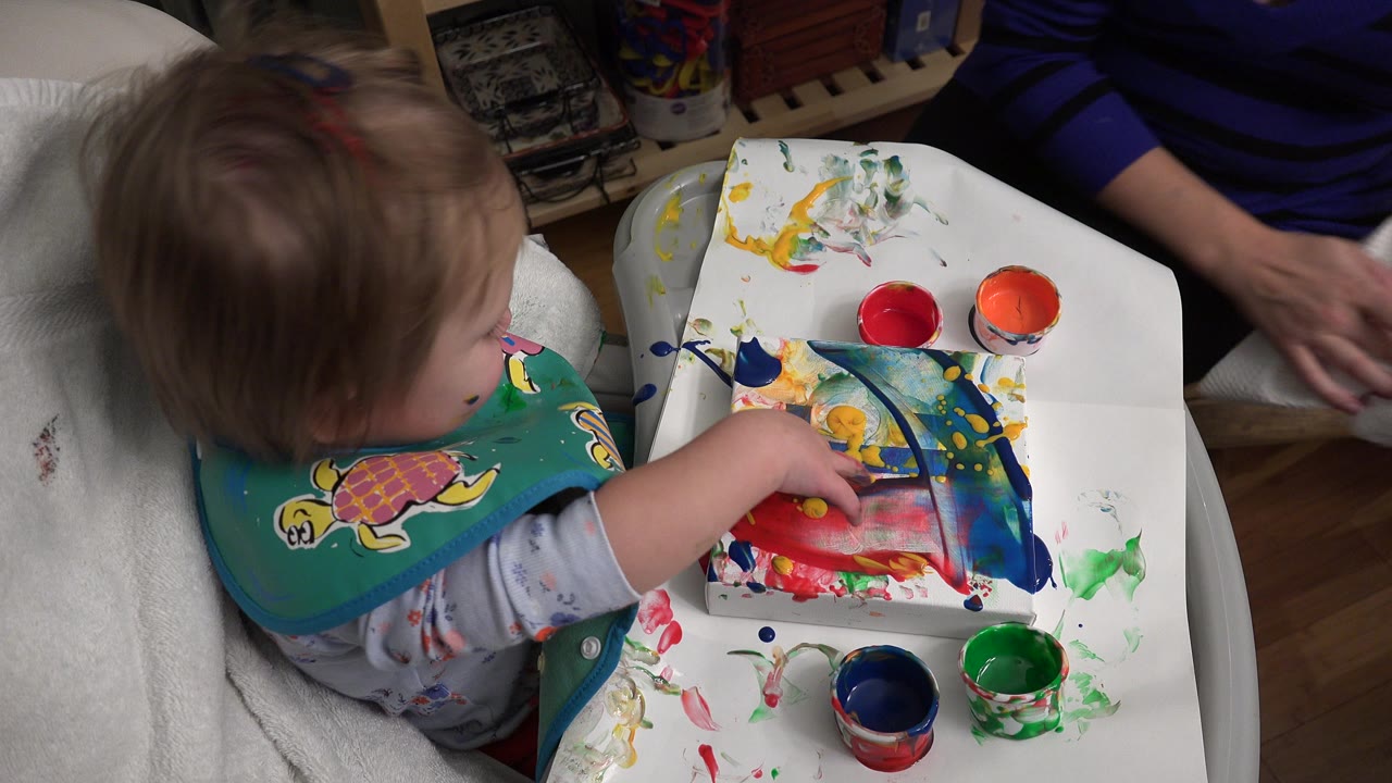 The toddler remains seated at the tray in Westminster, Colorado, with red, orange, blue, and green cups around a painted sheet while an adult's arm is visible nearby.