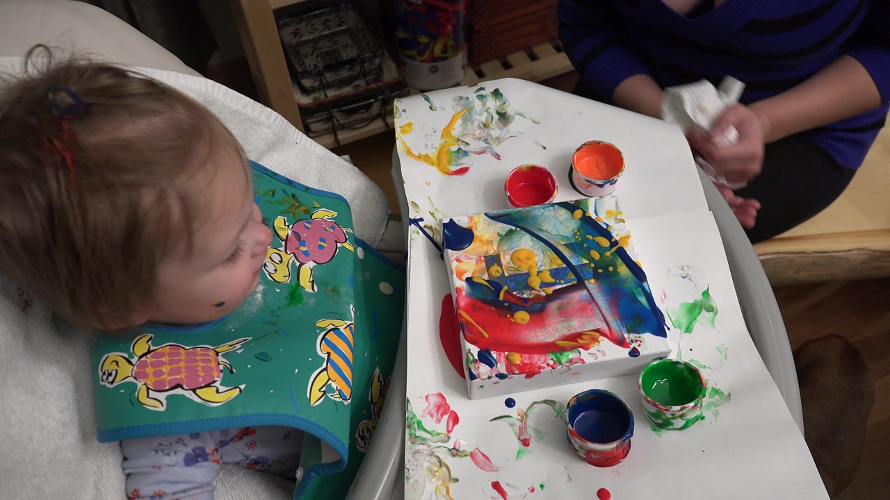 At 0:01 in Westminster, Colorado, the child looks across the high chair tray toward smeared paint and open cups as the adult's hands hold a cloth at the right edge.