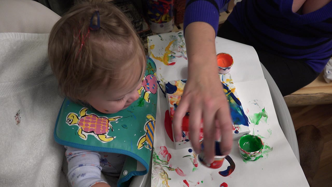 At 0:00 in Westminster, Colorado, a baby wearing a teal turtle bib sits in a high chair while an adult hand reaches toward paint cups on the tray.