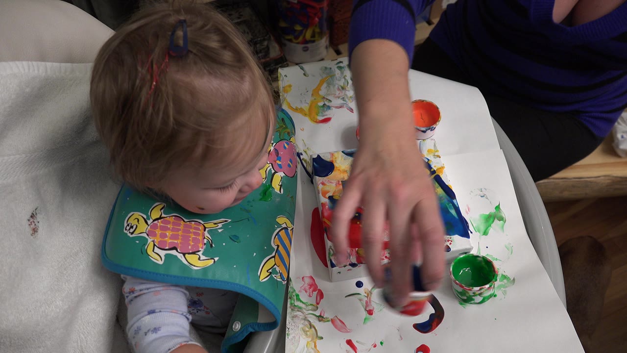 At 0:01 in Westminster, Colorado, the baby leans over the high-chair tray as an adult hand handles paint cups near smears of red, blue, and green color.