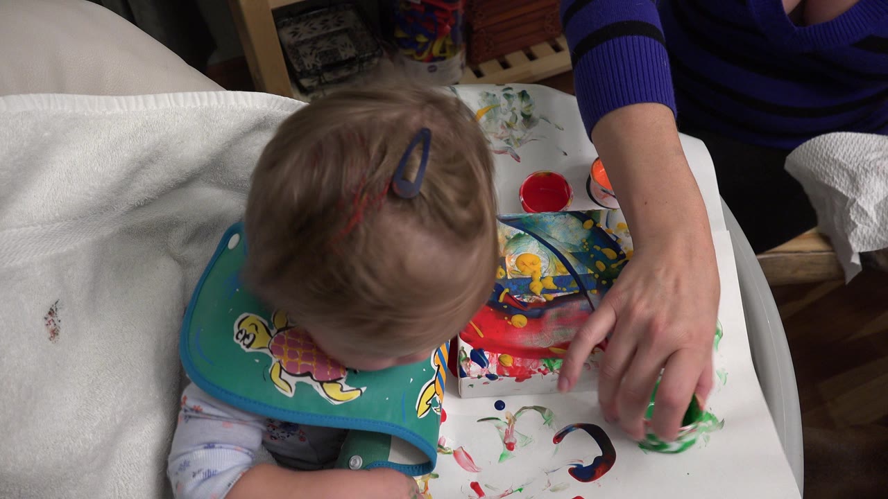 Toddler in a green bib sits beside colorful paint on a covered table.