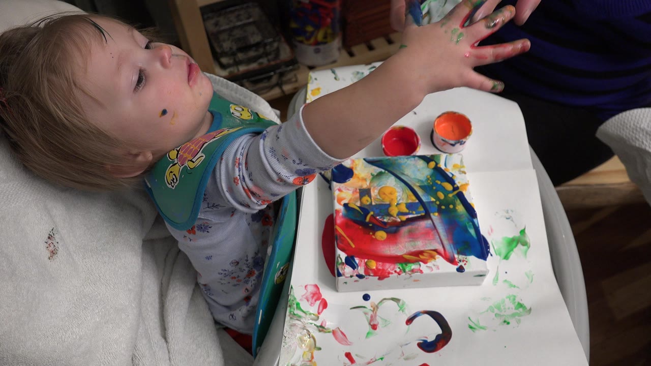 Baby in high chair reaches up beside a tray of colorful paints.