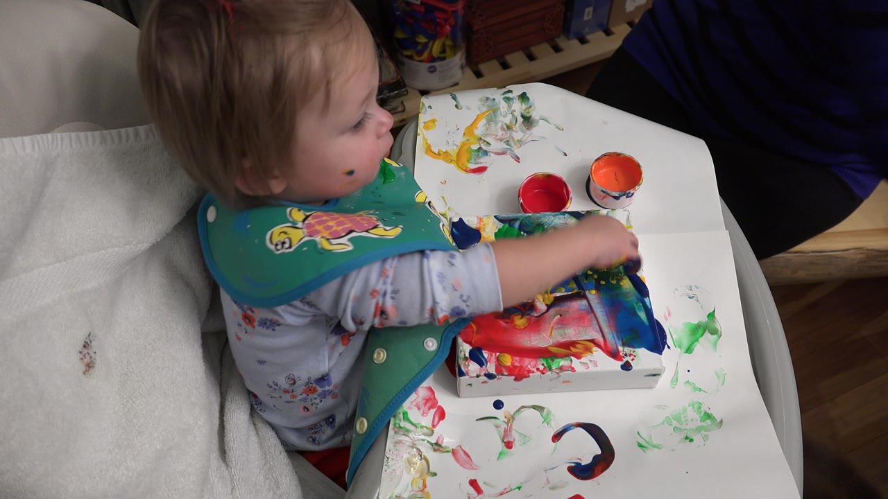 A baby girl in Westminster, Colorado sits in a high chair with a green turtle bib, reaching across a white tray covered in colorful paint and paper.