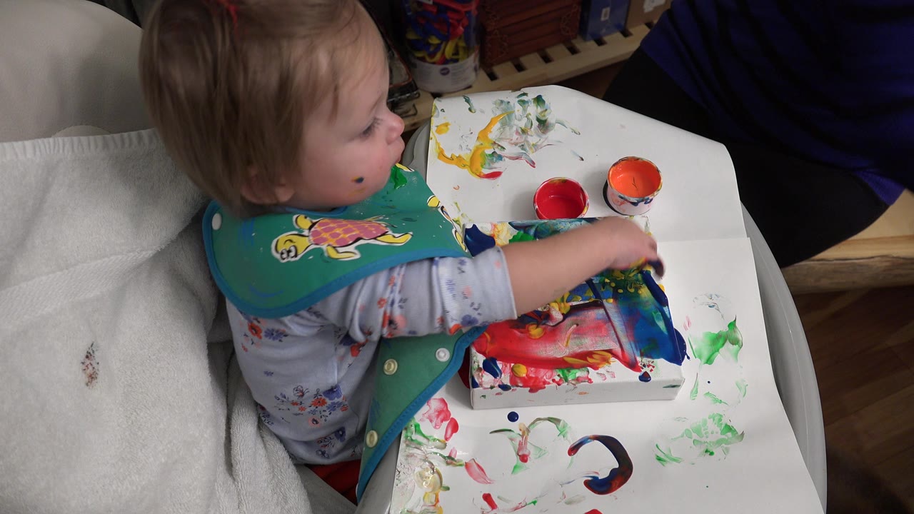 The baby girl remains seated beside red and orange paint cups, with her hand near a paint-smeared paper on the high-chair tray.