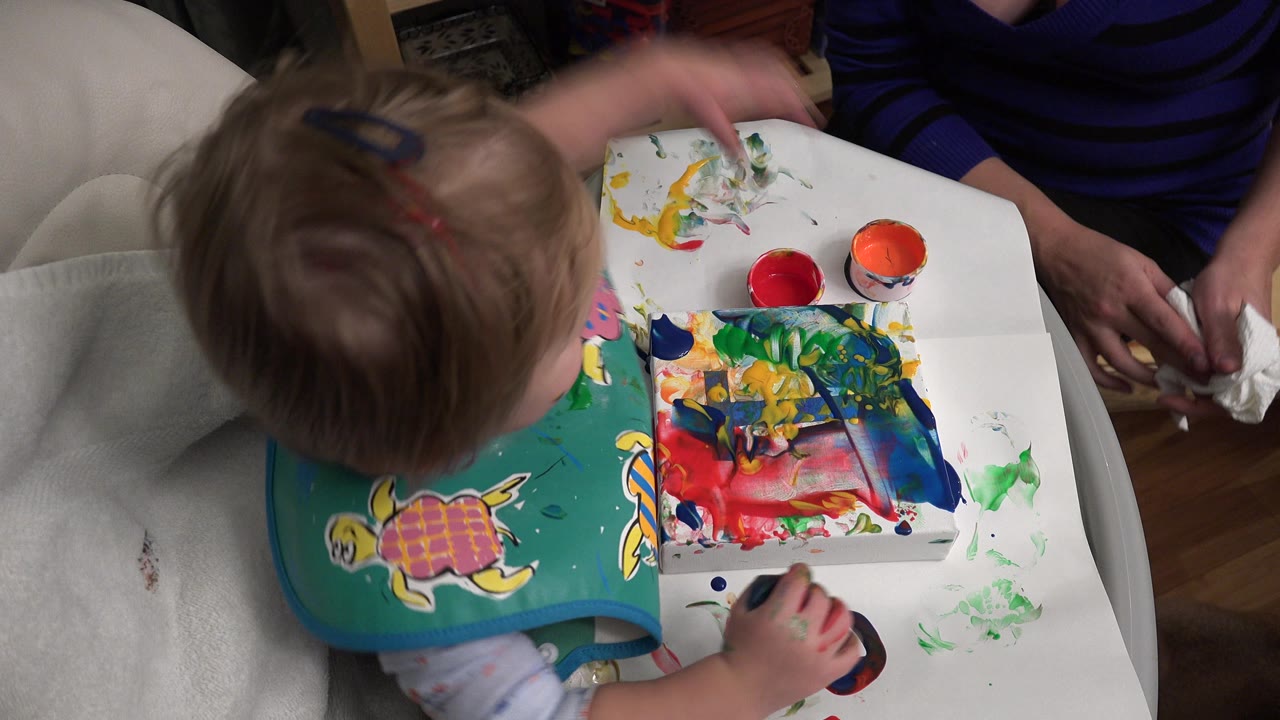 At 0:00 in Westminster, Colorado, a toddler wearing a green turtle bib reaches over a paint-smeared white table while an adult sits nearby with a cloth.