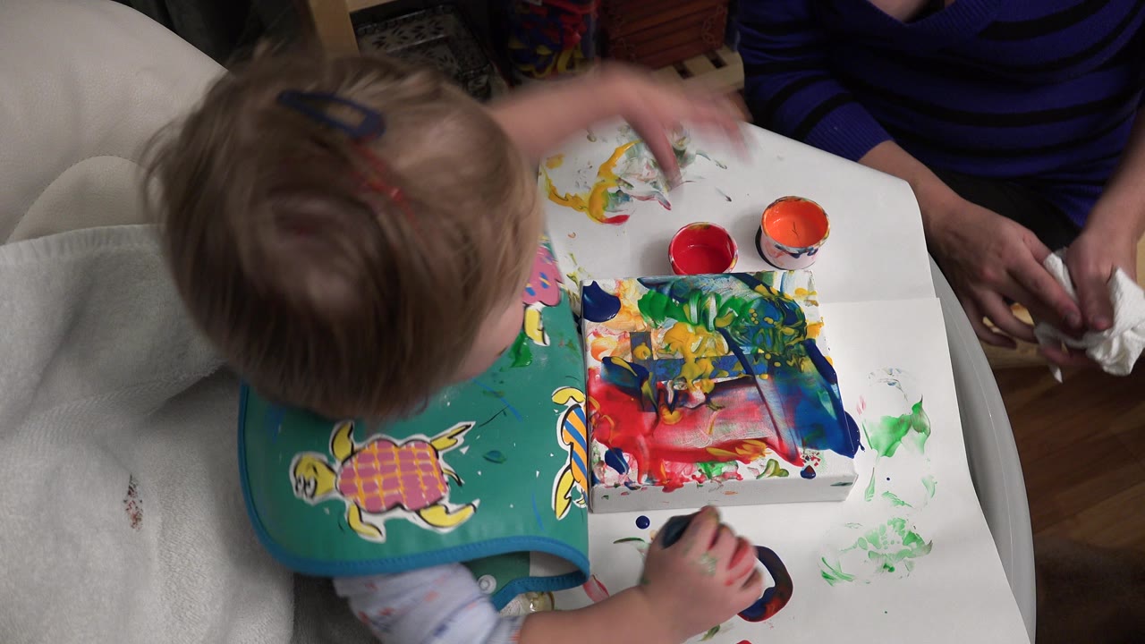 At 0:01 in Westminster, Colorado, the toddler leans over the table toward bright paint smears while the adult's hands remain visible beside a white cleaning cloth.