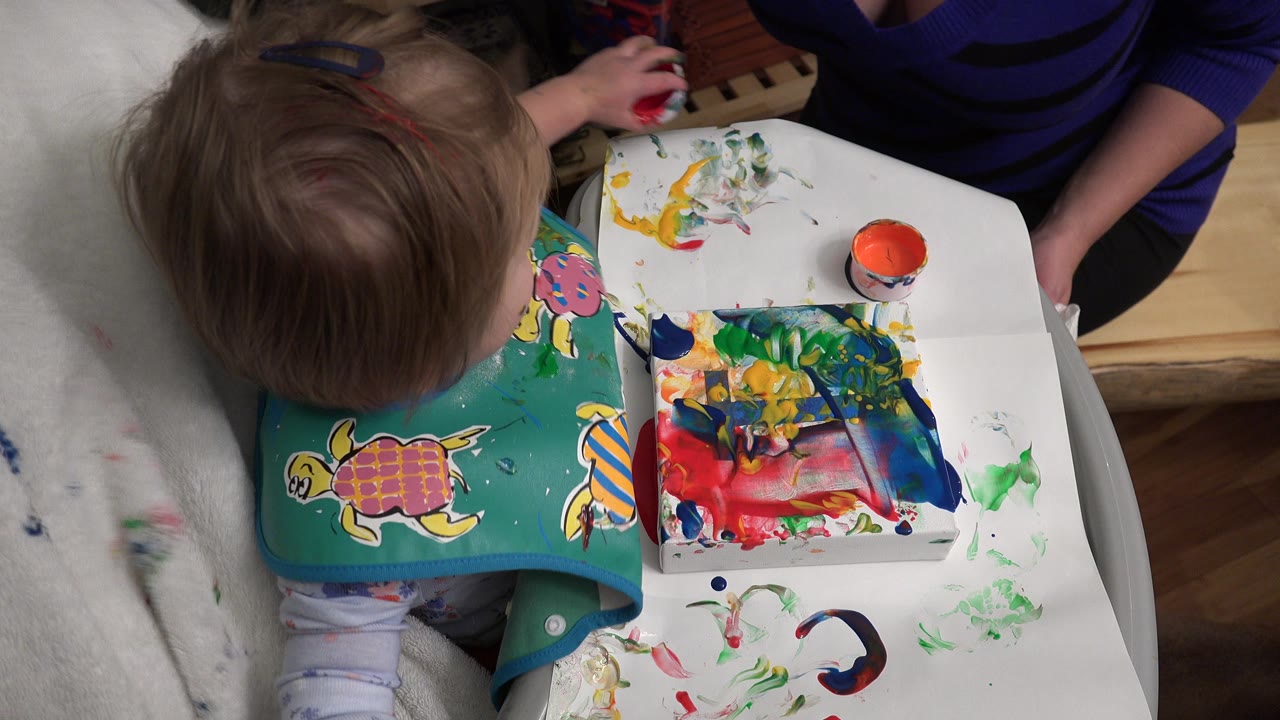 Baby at a small table with paint cups and a colorful painting.