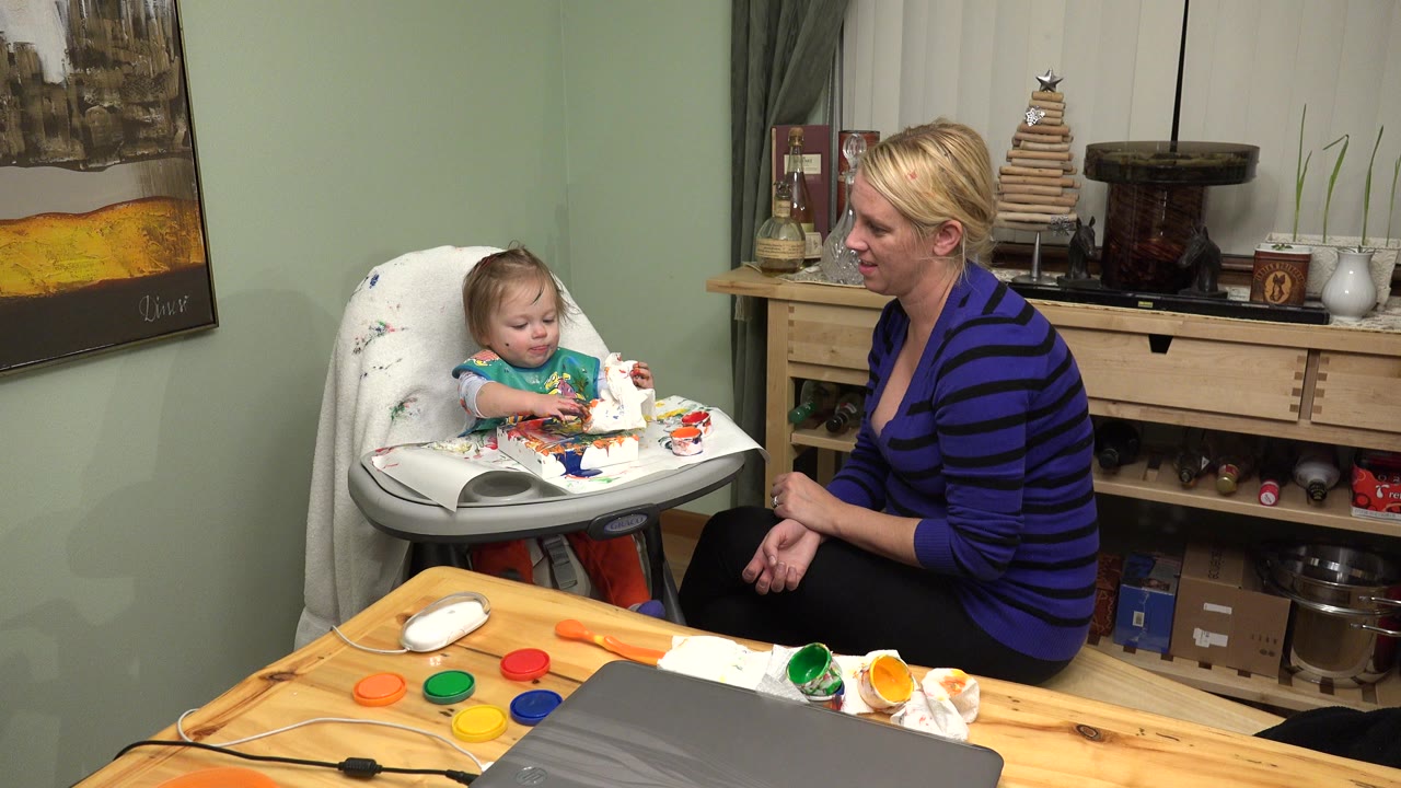 In Westminster, Colorado, a baby sits in a high chair with paint on the tray while an older blonde woman watches from beside the chair at the start of the clip.