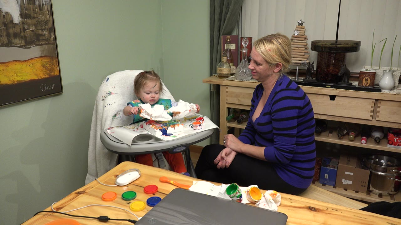 In Westminster, Colorado, the baby looks down at the cluttered highchair tray as the woman watches from the right side of the room.