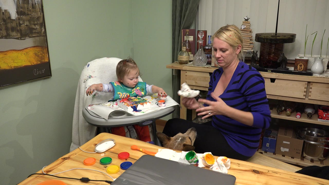 In a Westminster, Colorado home interior, a mother sits at a wooden table while a young girl in a high chair looks at colorful art materials spread across the tray.