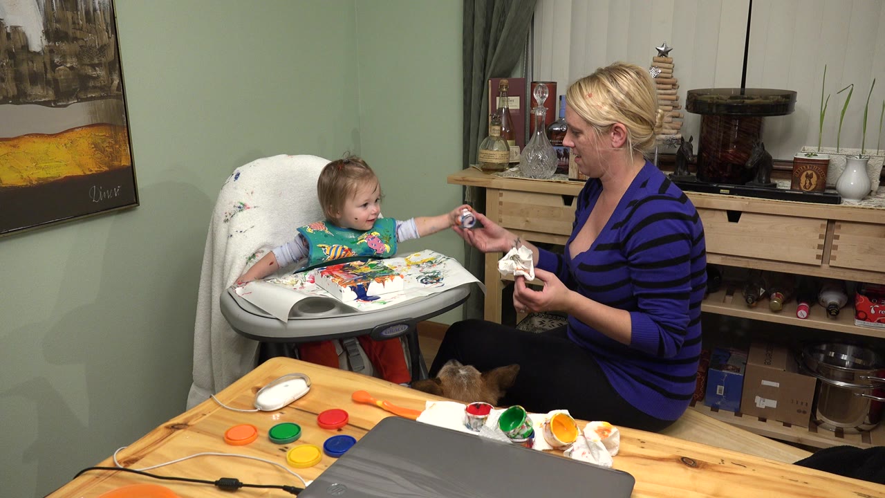 Woman offering paint to a baby seated in a high chair.