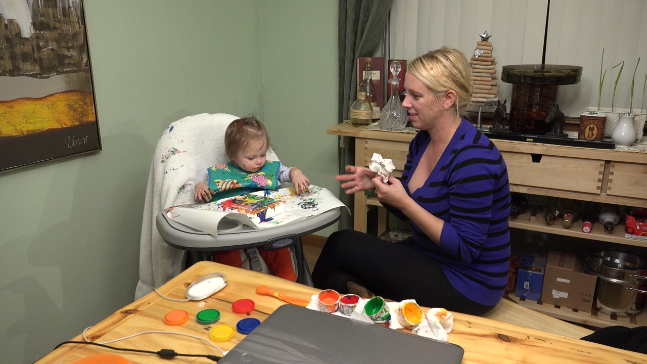 In Westminster, Colorado, a mother sits beside a toddler in a high chair as the child works with bright paint on paper at an indoor table.