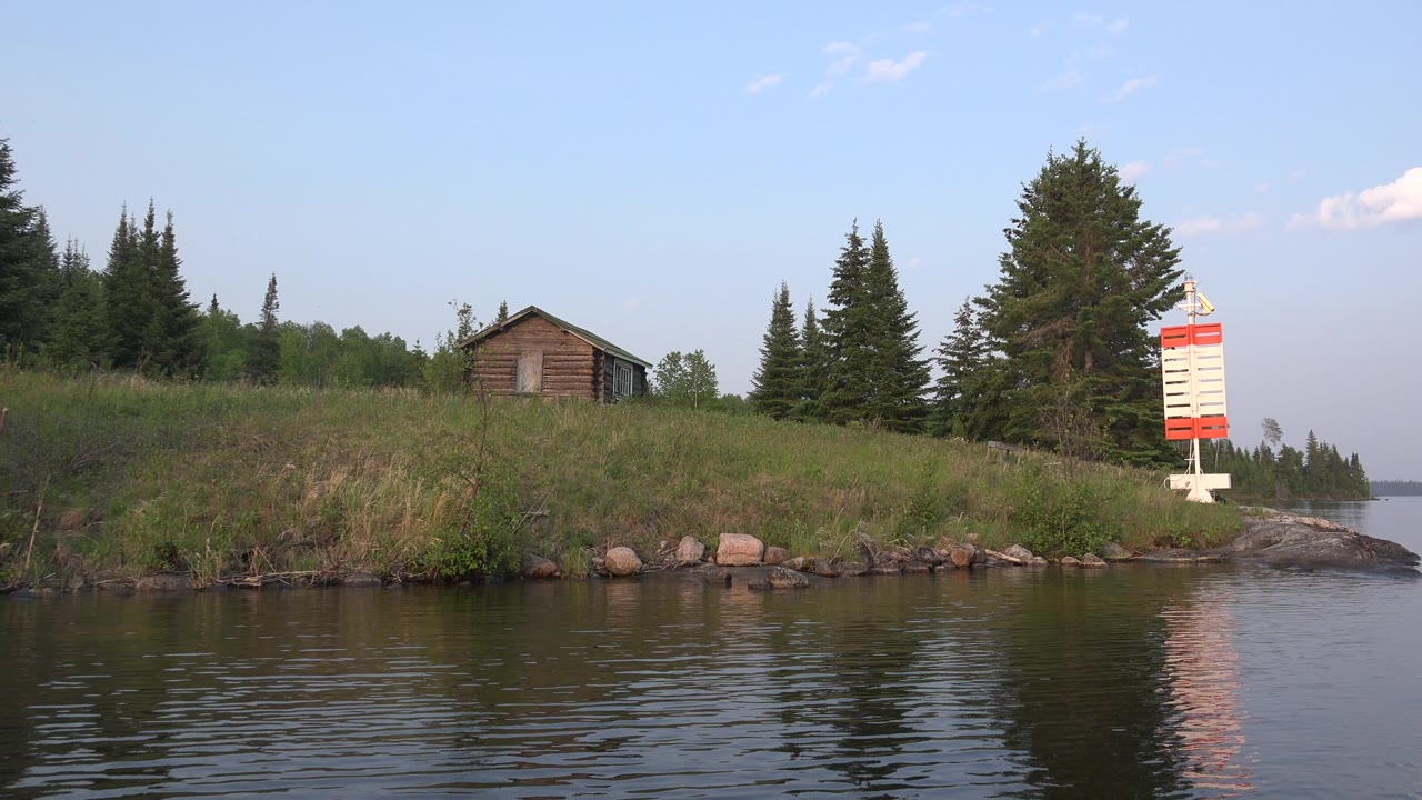 Red Lake, Canada frame with the cabin left of center, dark evergreen trees along the bank, water ripples across the lower frame, the red-and-white marker near the right edge, and no visible people.