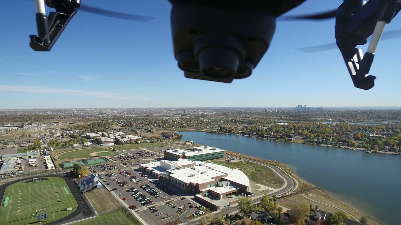 Second aerial frame in Hidden Lake, Colorado, showing the drone hardware at the top, sports fields and parking below, water across the middle, and no visible people.