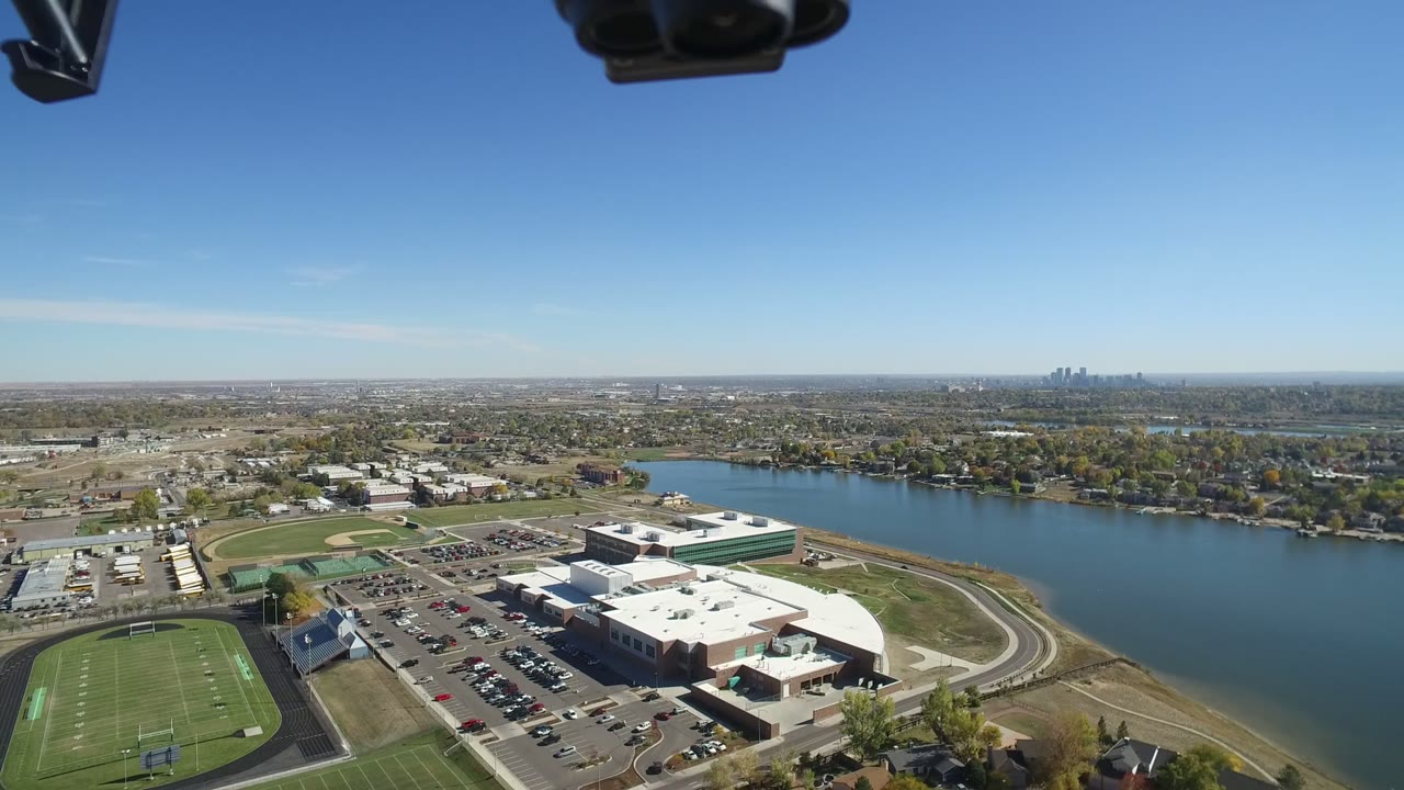Aerial view of sports fields, large buildings, parking lots, a lake, and distant skyline.