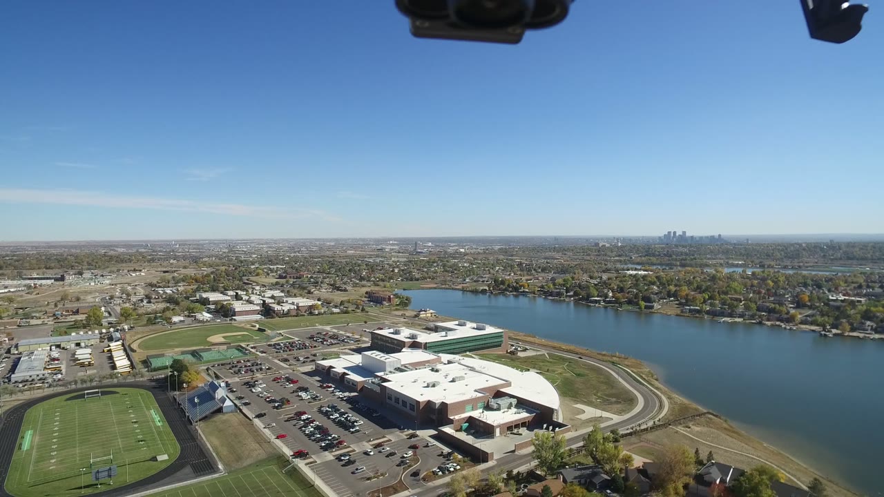 Second aerial frame in Hidden Lake, Colorado, showing the sports fields and parking lot slightly more centered, the lake stretching across the midground, low neighborhoods beyond, and no visible people.