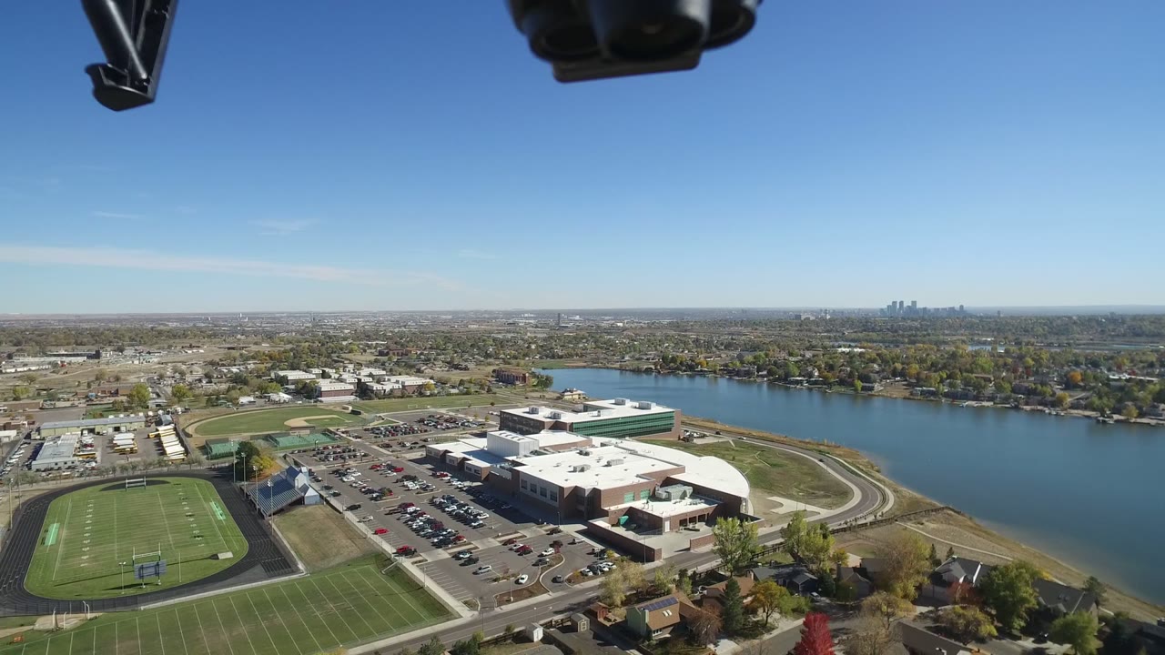 Aerial view of sports fields, a large white building, parking lots, water, and distant suburbs.