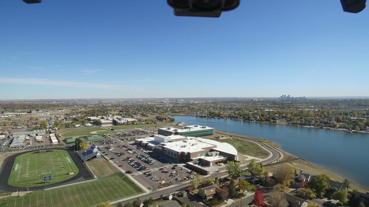 One second into the Hidden Lake, Colorado aerial, the frame holds athletic fields at left, parked cars and a large building near center, water behind them, and no visible people.
