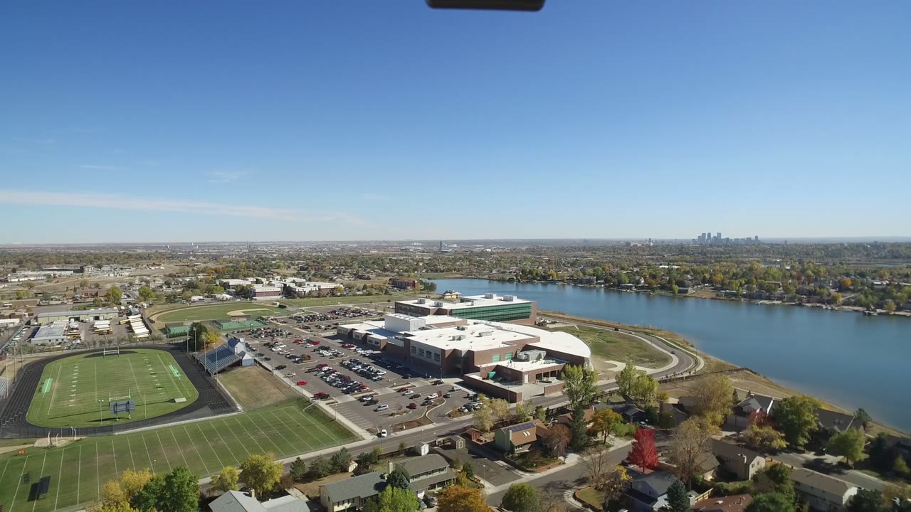 Wide aerial frame over Hidden Lake, Colorado showing a football field at left, a large school building and parking lots near the water, homes in the foreground, and no visible people.