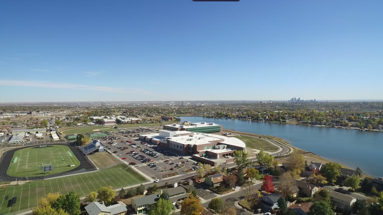 Aerial view in Hidden Lake, Colorado with the athletic field prominent on the left, the school complex centered near parked cars, a wide waterway behind it, and no visible people.