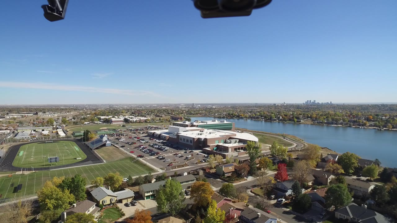 Aerial view of homes, trees, school field, parking lots, and water under blue sky.