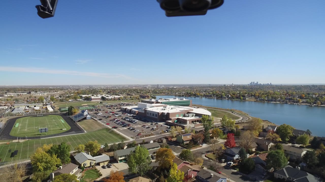 Second aerial frame in Hidden Lake, Colorado, with the sports field on the left, parking lots and large buildings centered, residential roofs below, water across the upper right, and no visible people.