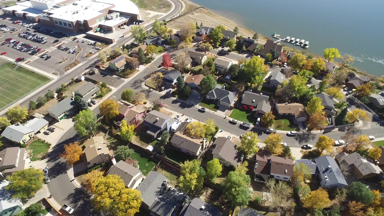 Oblique drone frame over Hidden Lake, Colorado rooftops, autumn trees, parked cars, rectangular sports fields, lake shoreline, and no visible people.