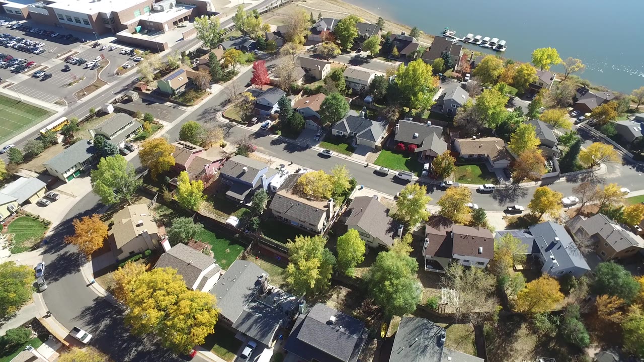 Aerial view of homes, roads, trees, parked cars, and lake edge.