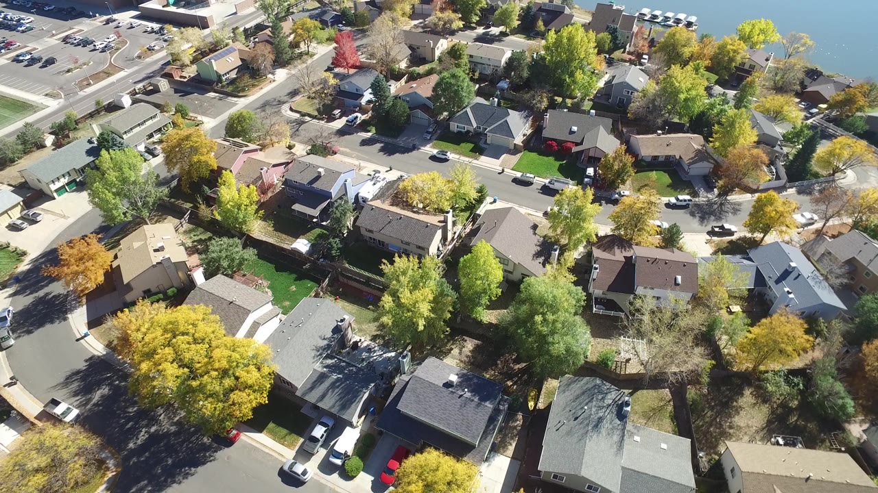 High overhead view in Hidden Lake, Colorado with houses, tree-lined streets, parked cars, and a small lake edge near the upper right, with no visible people.
