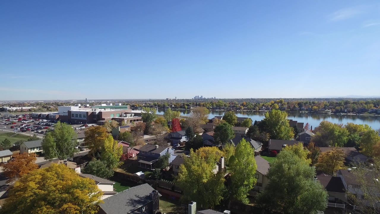 Opening aerial frame over Hidden Lake, Colorado with rooftops and autumn trees in the foreground, a parking lot and lake beyond, distant skyline on the horizon, and no visible people.