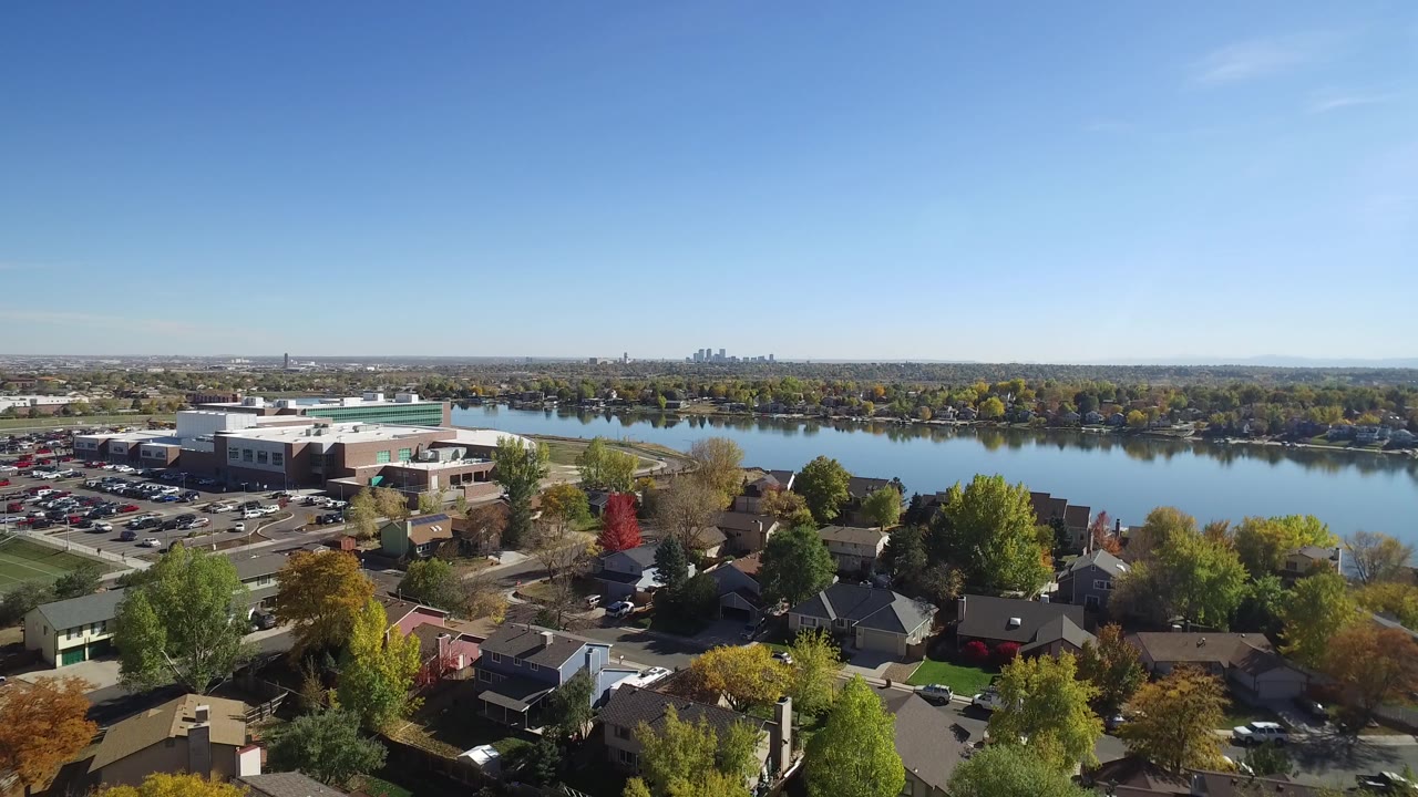 Aerial view of a lake beside houses and autumn trees under a clear sky.