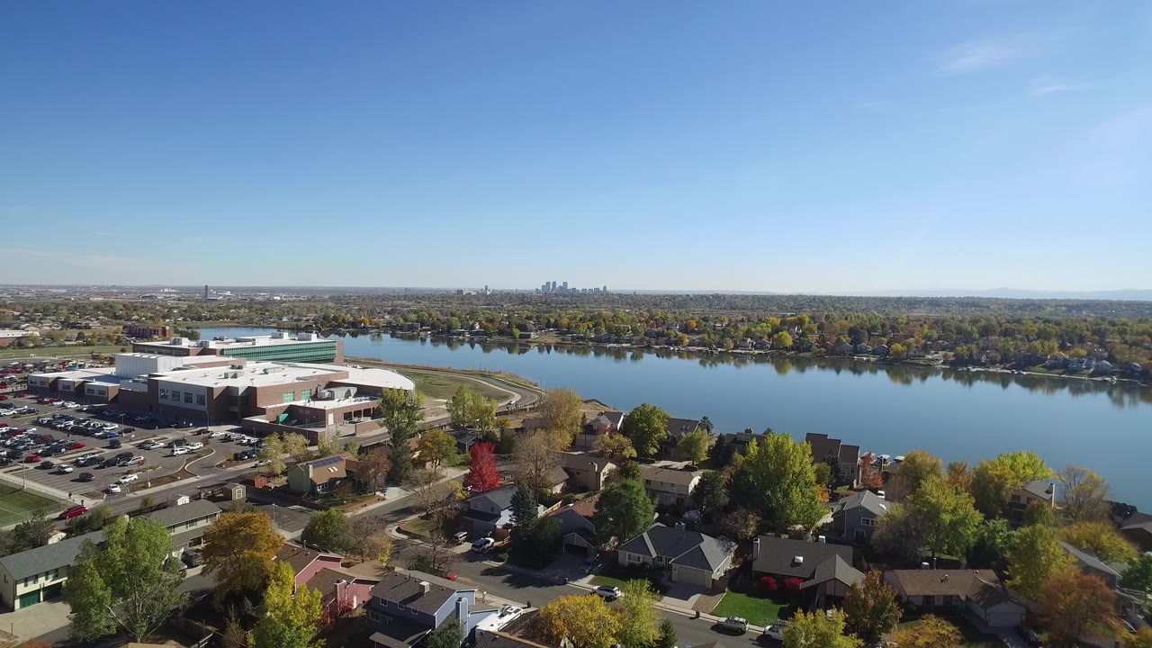 High aerial view over Hidden Lake, Colorado with homes in the foreground, large buildings and parking lots at left, a lake across the middle, distant skyline, and no visible people.
