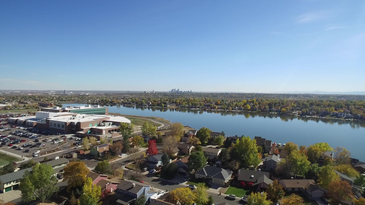 Aerial suburban lake scene with flat-roofed buildings, houses, trees, and blue sky.