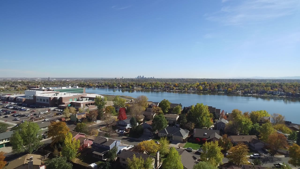 Aerial frame over Hidden Lake, Colorado showing rooftops, a large light-roofed building, a broad lake, autumn trees, distant skyline, and no visible people.