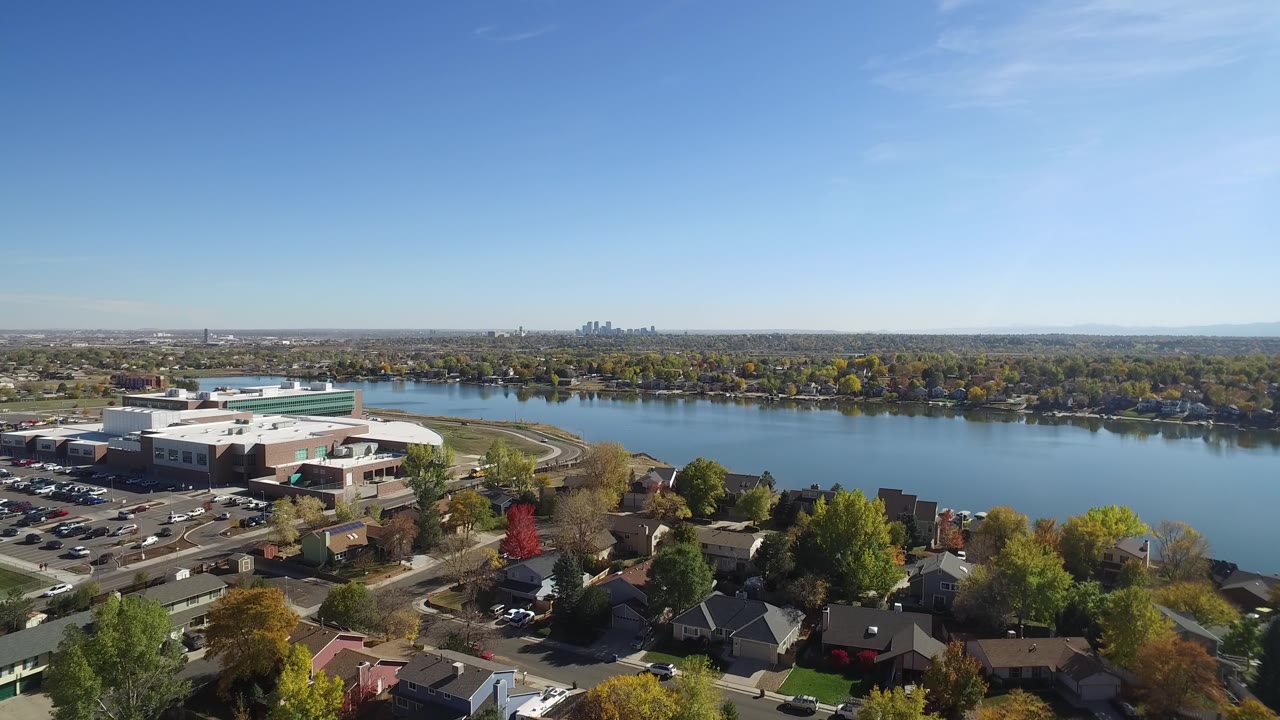 At 0:01, the Hidden Lake, Colorado view holds on the white building and parking area at left, blue water across the center, houses below, distant skyline on the horizon, and no visible people.